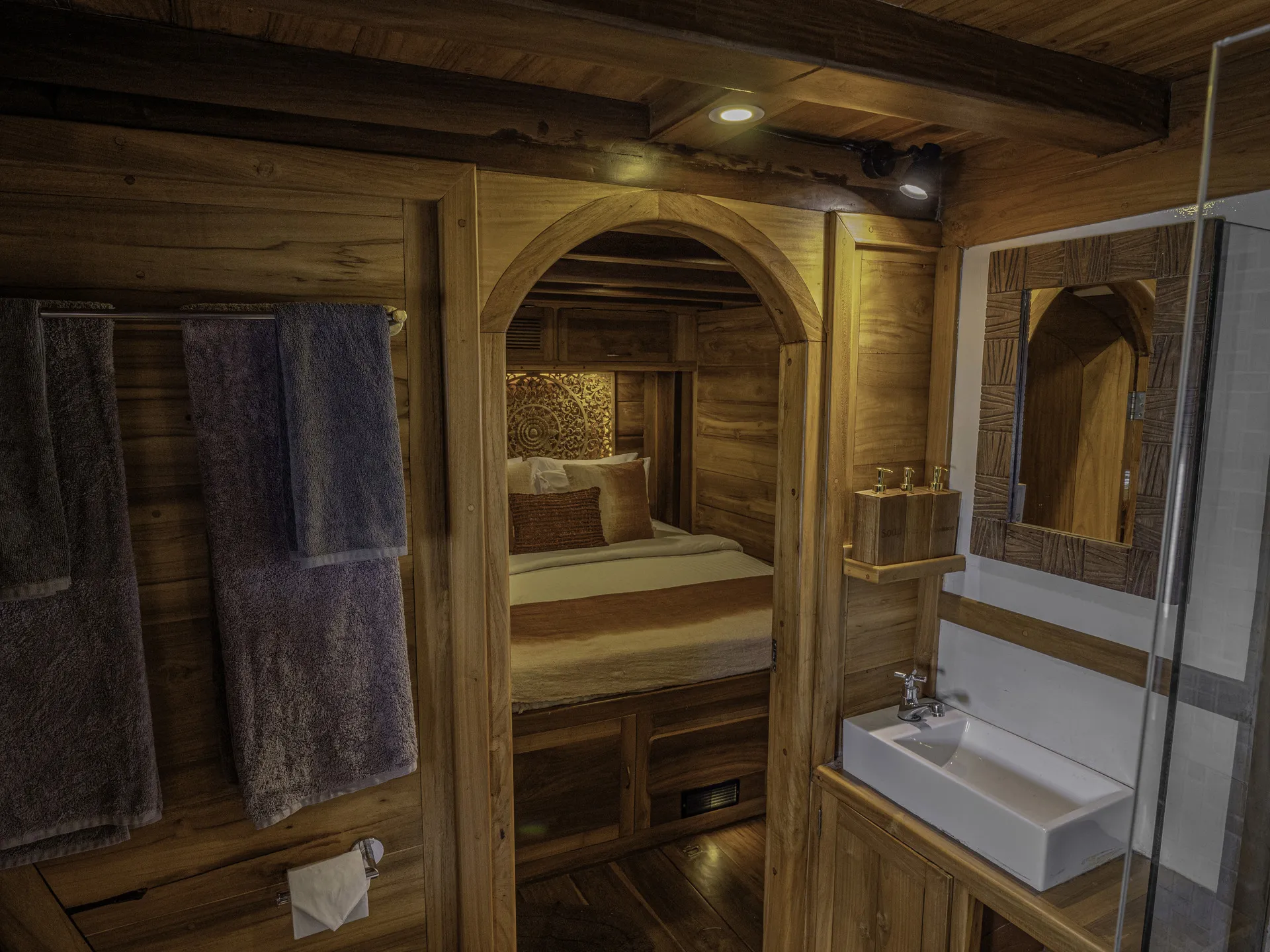 A cozy wooden cabin bathroom, reminiscent of a charter yacht in Indonesia, features a white sink, mirror, glass shower, and shelves. Through an arched doorway, a neatly made bed with pillows and rustic wood walls is visible. Two towels hang on a rack.