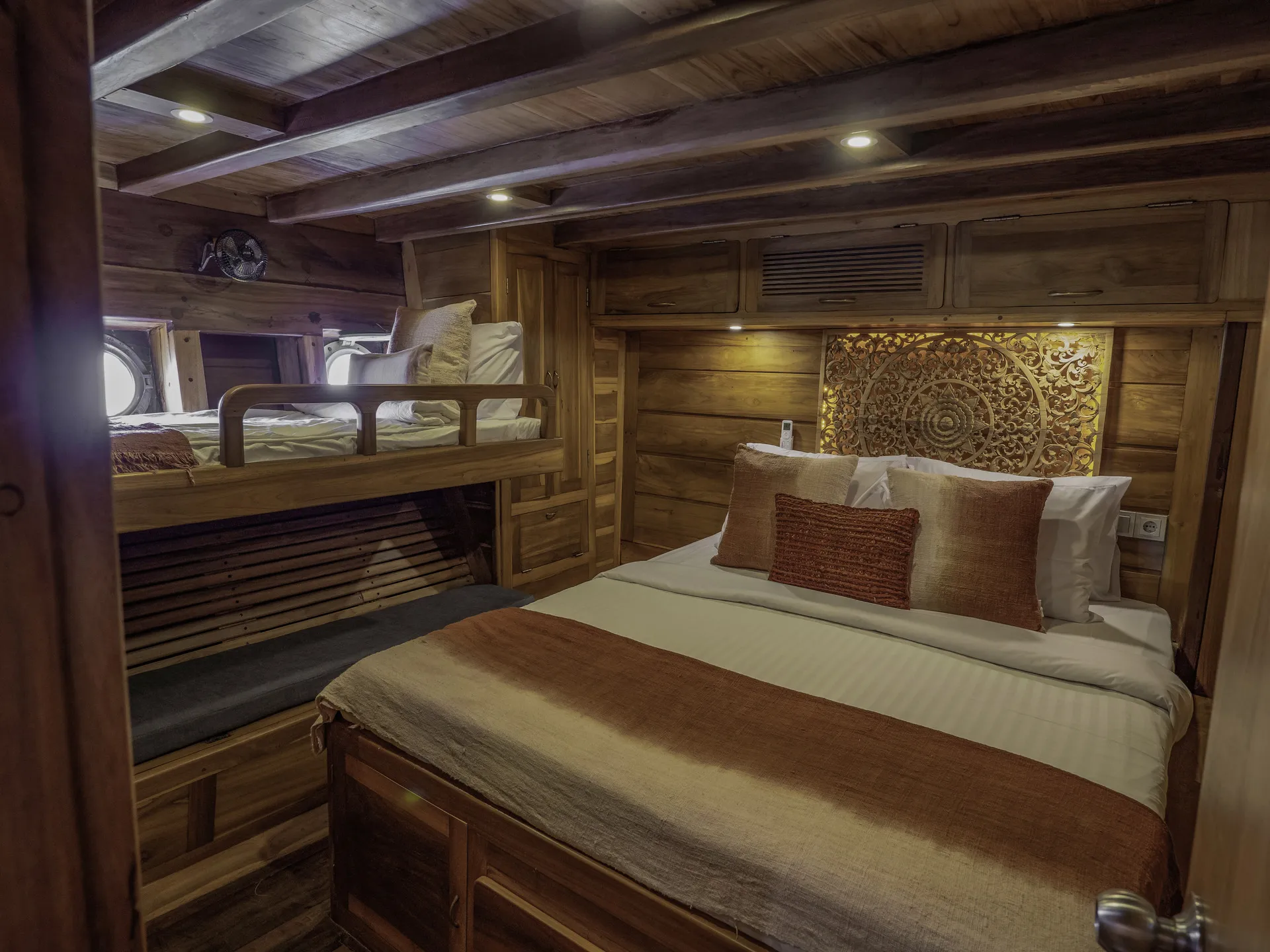 A cozy wooden cabin bedroom on a charter yacht, with a double bed and bunk beds, accented with white and brown bedding, decorative pillows, and an Indonesian carved wooden wall panel, warmly lit by ceiling lights.