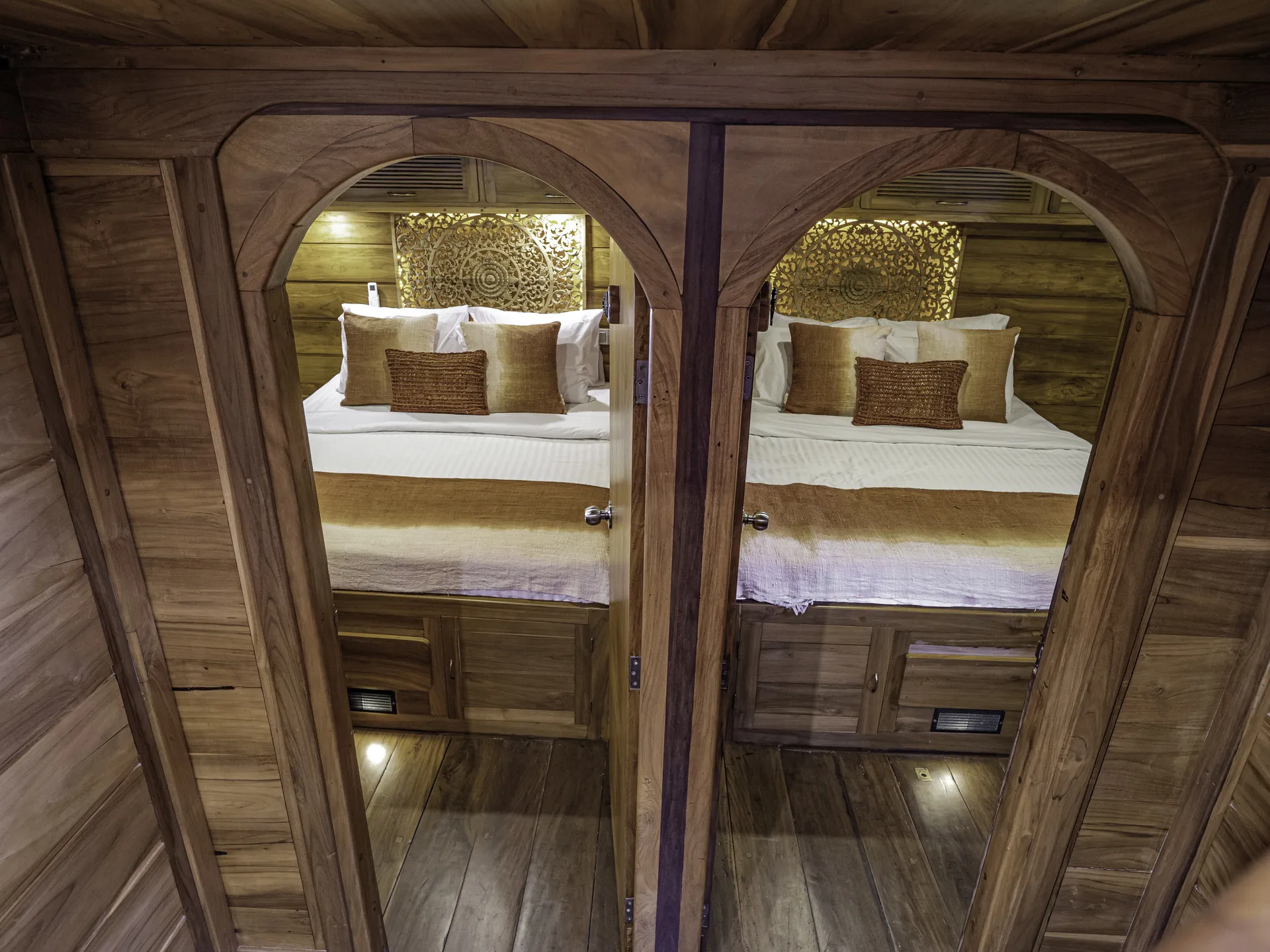 Two adjacent cozy beds with white sheets and decorative brown pillows, separated by a wooden partition with arched doorways, set in a warmly lit, wood-paneled room reminiscent of a charter yacht retreat in Indonesia.