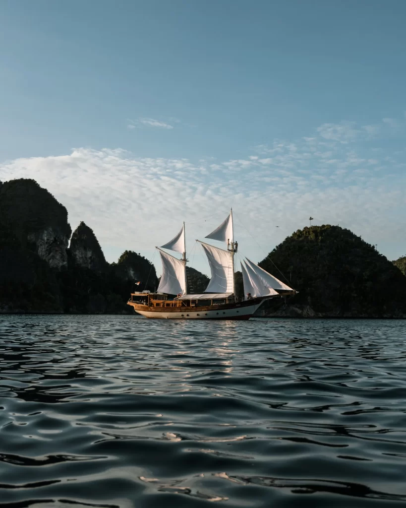 A large charter yacht with white sails glides on calm water, surrounded by green, rocky islands in Indonesia under a clear sky with scattered clouds.