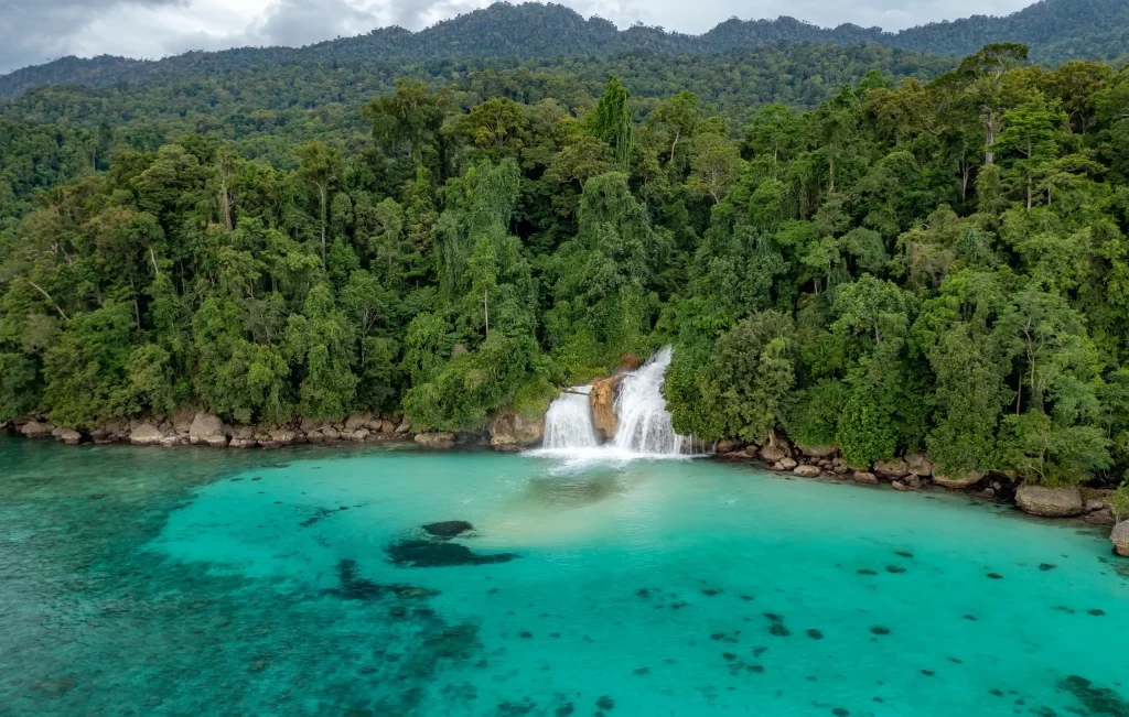 Stunning tropical waterfall flowing directly into the turquoise sea in Triton Bay, Indonesia, surrounded by lush rainforest and pristine waters.
