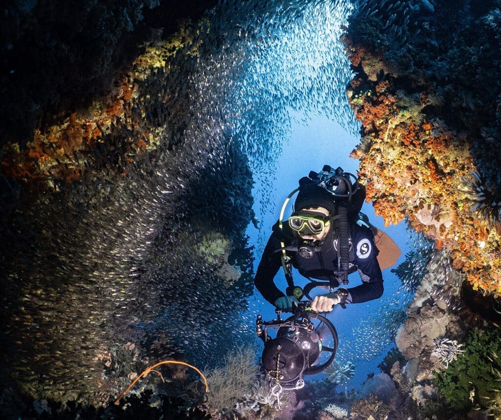 Scuba diver exploring a vibrant coral reef cave surrounded by a school of fish in Raja Ampat, Indonesia, a top liveaboard diving destination.