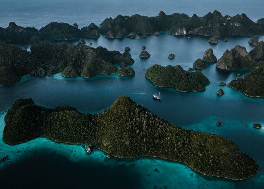 Stunning aerial view of Raja Ampat's lush green islands surrounded by turquoise waters, featuring a traditional yacht sailing through the serene sea.
