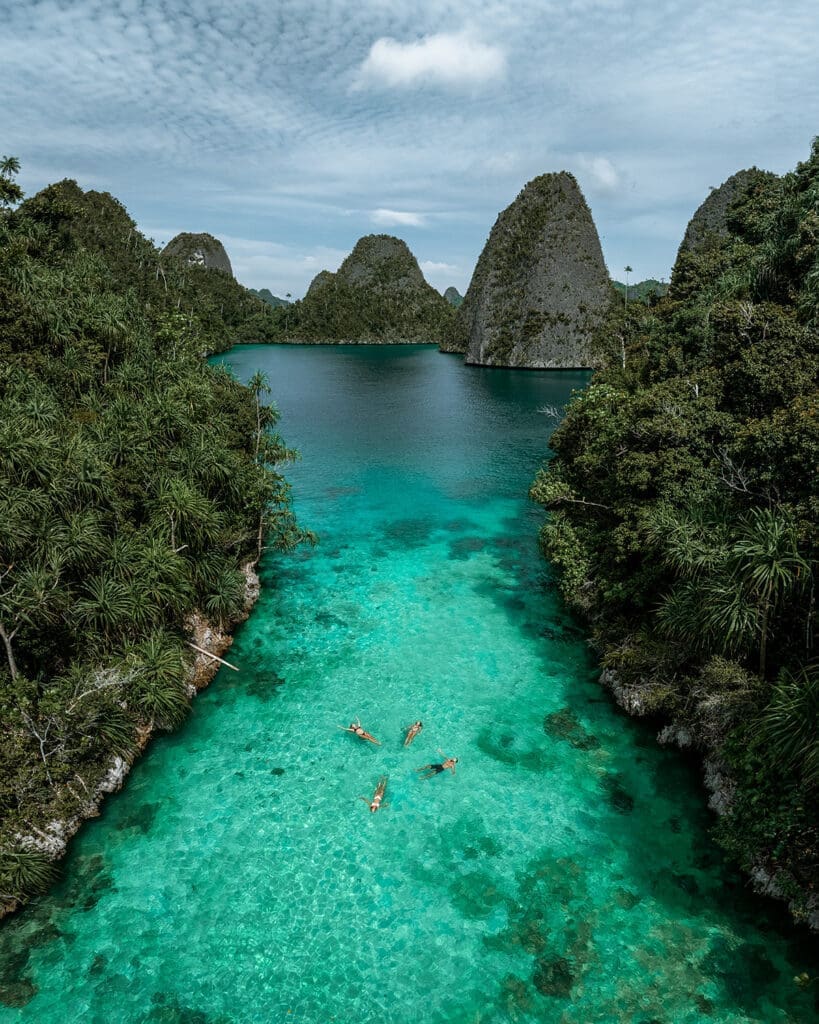 Aerial view of travelers swimming in the crystal-clear turquoise waters of Wayag, Raja Ampat, surrounded by lush greenery and towering limestone karst formations—an iconic destination for yachting, snorkeling, and exploration.