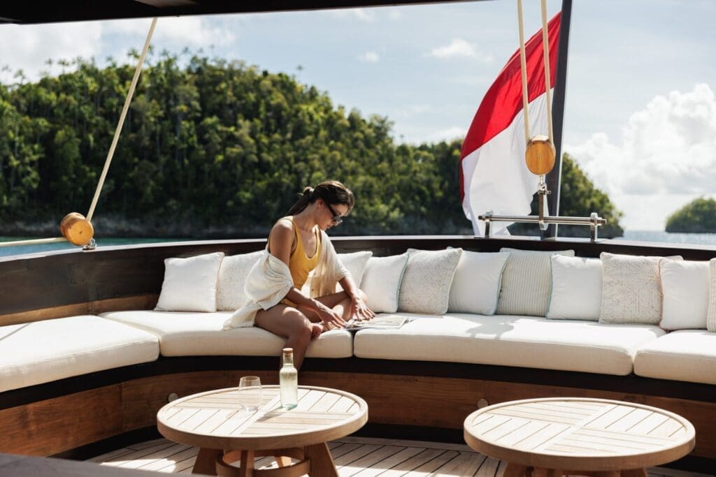 a young lady sitting on a soft white sofa reading a magazine at the back of a boat.